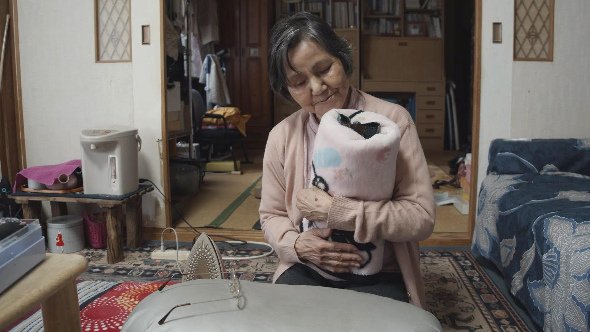 an elder woman sitting in her home environment, holding and looking at a wrapped blanket as if it's a baby